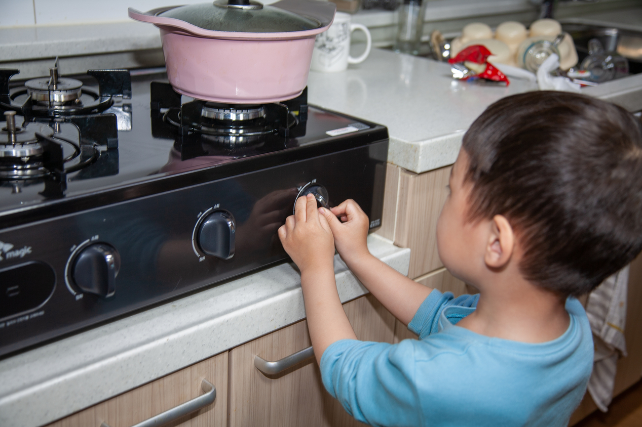A small child standing in front of a gas stove, with both hands adjusting the burner controls and a pot on the front right burner. 