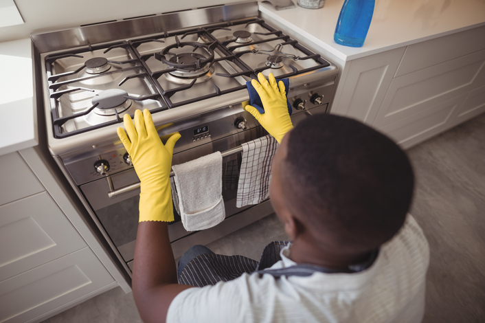 A person crouching down and cleaning their gas stove. They are wearing yellow gloves and have a scrubber in their right hand. 